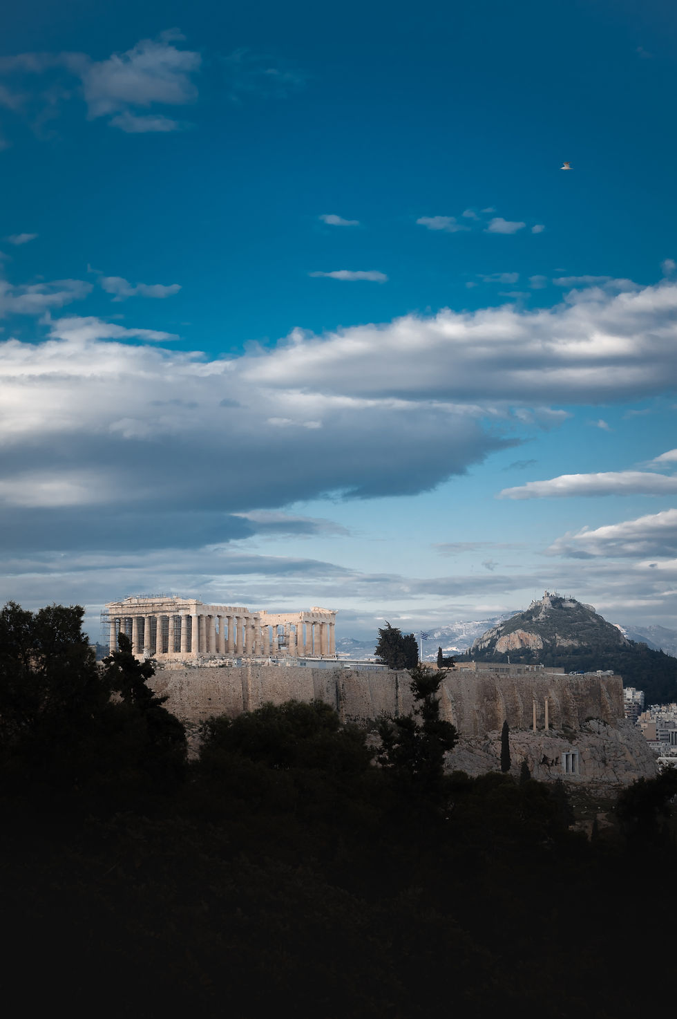 The Parthenon standing atop the ancient Acropolis in Athens, Greece, with Mount Lycabettus visible in the background under a partly cloudy sky.