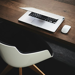 Halbgeöffneter silberner Laptop und weiße Computermaus auf einem Holztisch, davor ein weißer Stuhl mit schwarzer Polsterung.