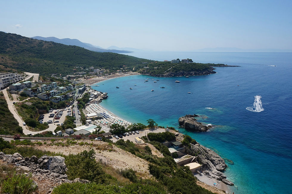 Coastal view with turquoise sea, boats, and beach umbrellas. Green hills and buildings line the shore under a clear blue sky.