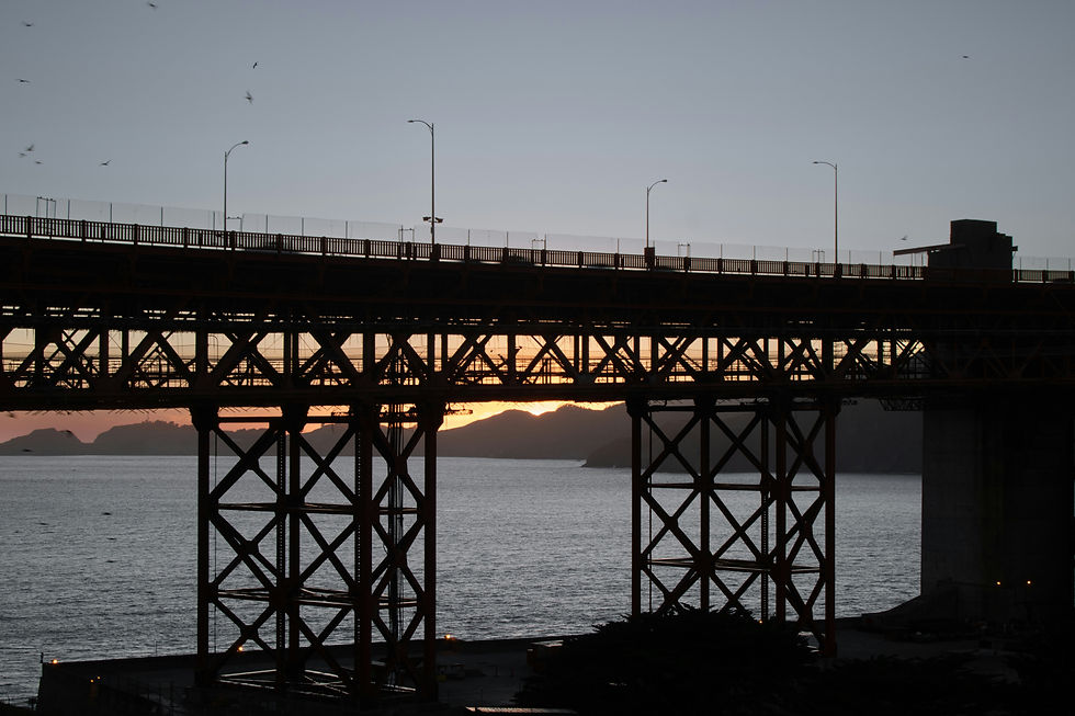 The image shows a bridge structure silhouetted against a setting sun, casting a dramatic shadow over the calm water below. The steel framework and supporting pillars are clearly visible, adding an industrial
