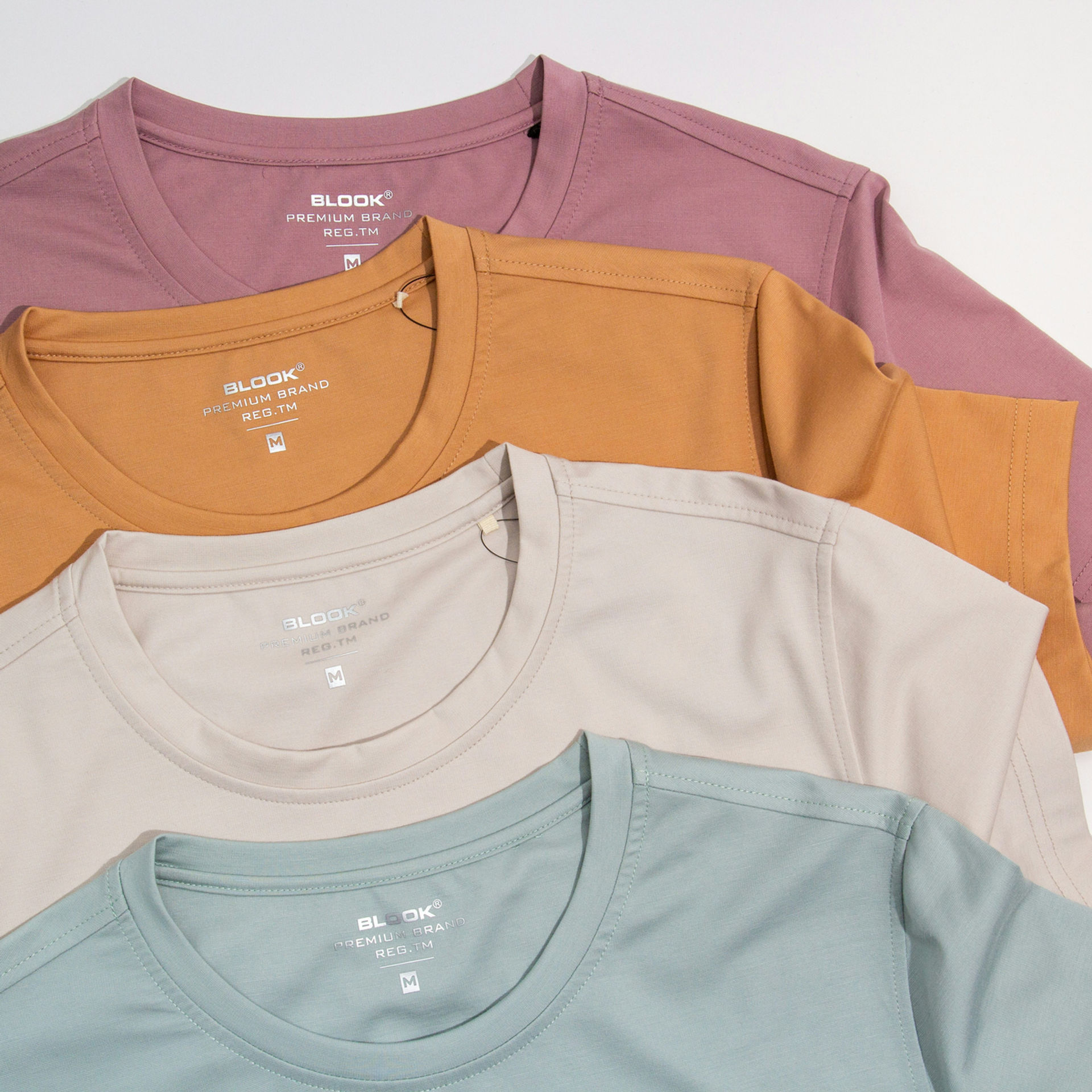 Four neatly stacked T-shirts in pink, mustard, beige, and teal. Each shirt has "BLOOK PREMIUM BRAND" printed on the chest.