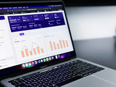 Laptop displaying financial dashboard with graphs and metrics on screen. Purple and white interface, desk background, calm setting.