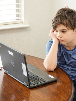 Boy in blue shirt looks bored at a table with a laptop, paper, and pencil. Bright window blinds in the background.