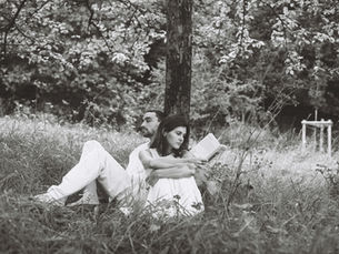 Black-and-white photo of a couple sitting back-to-back by a tree in tall grass, the woman reading a book in a calm forest.