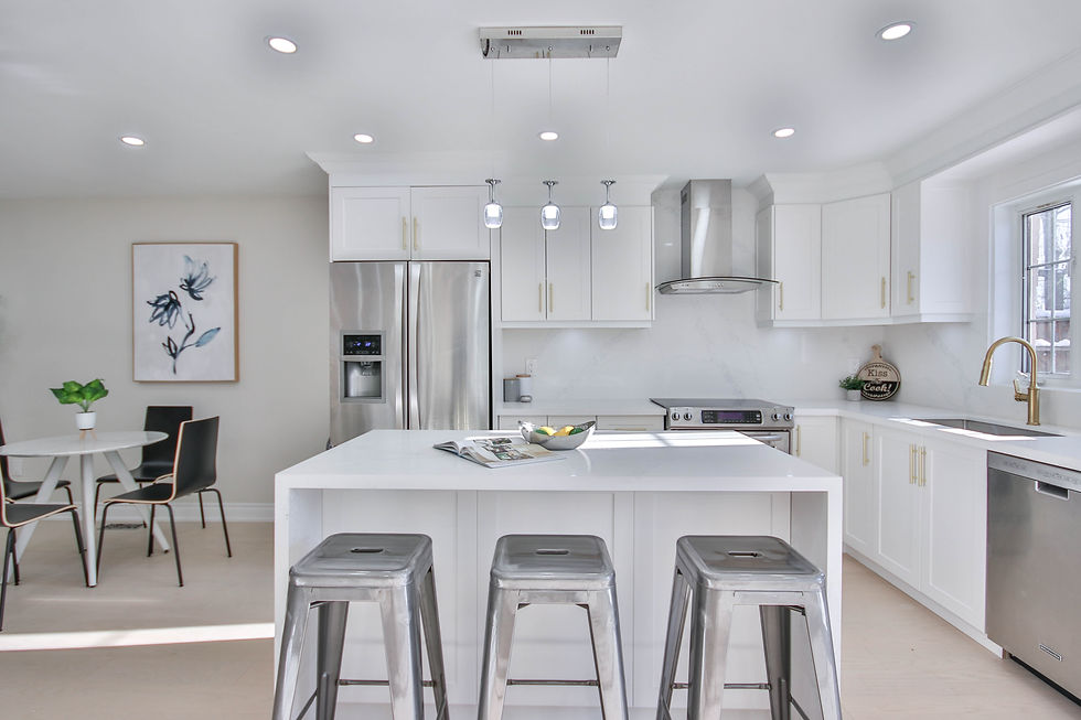 Modern kitchen with white cabinets, stainless appliances, and a large island with stools. A small dining table and plant are visible. Bright and airy.