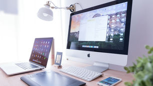 Moderner Arbeitsplatz mit iMac, MacBook, Tablet und Smartphone auf einem hellen Schreibtisch, ausgestattet mit einer Schreibtischlampe und Grafiktablett.