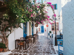 Charming Greek alley with cobblestone path, lined with tables, blue doors, and pink flowers. Sunlit, peaceful, Mediterranean vibe.
