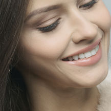 Close-up of a smiling woman with closed eyes, dark hair, and glossy lips against a soft light background.