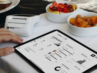 En hånd betjener et nettbrett med salgstall og analyser, ved siden av en kortterminal og skåler med snacks – et visuelt eksempel på hvordan småbedrifter kan bruke data og Google Ads for småbedrifter til å ta bedre markedsføringsbeslutninger.