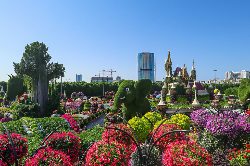 Colorful garden with floral sculptures of an elephant and castle, surrounded by vibrant flowers. Skyscraper and clear blue sky in background.