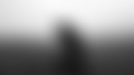A person in dark clothing sits on an empty road, head in hand, looking despondent. Foggy background with faint outlines of road signs. Black and white.