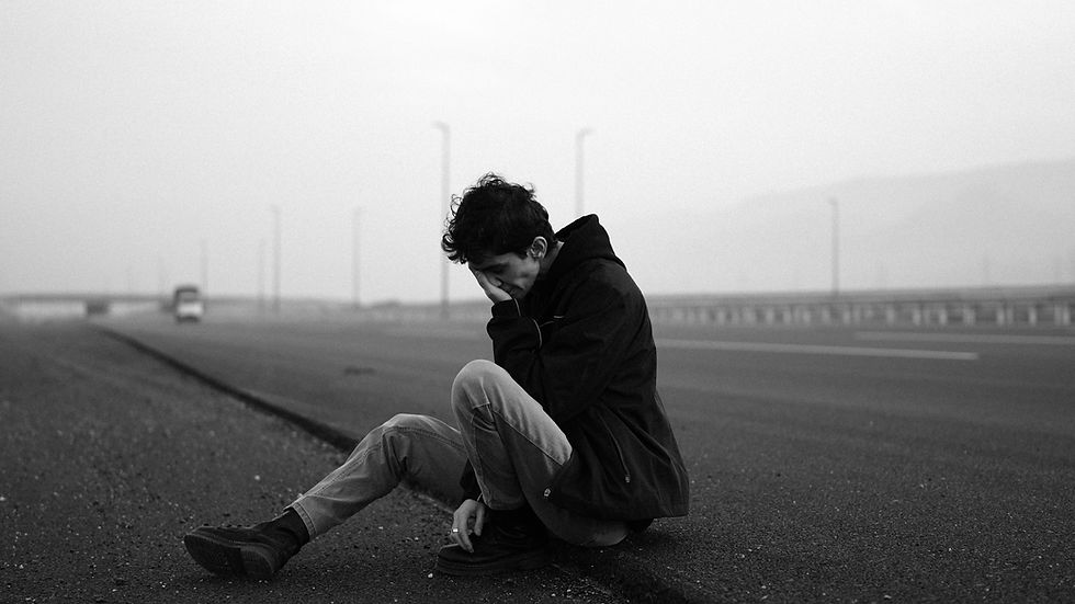 A person in dark clothing sits on an empty road, head in hand, looking despondent. Foggy background with faint outlines of road signs. Black and white.