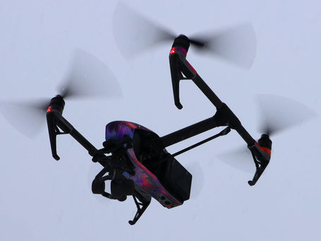 A colorful drone flies against a gray sky with spinning propellers visible. Red lights glow on the drone's arms.