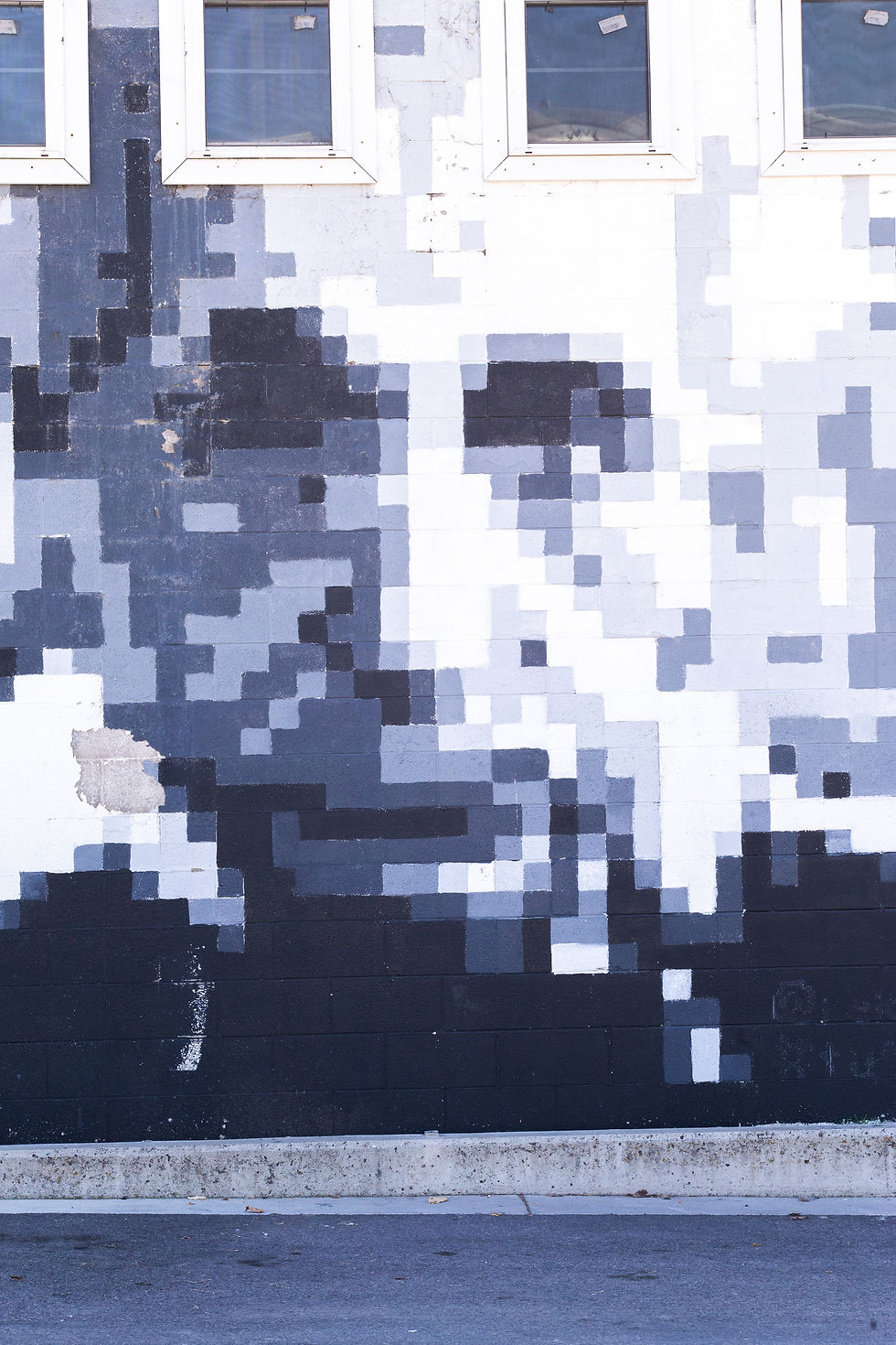 Pixelated black-and-white mural of a face on a brick wall, under three square windows. Urban street setting with concrete curb.