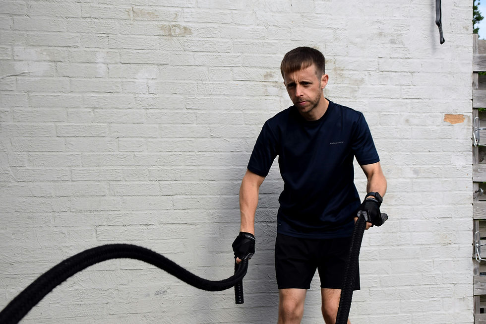 Homme en tenue de sport noire s’entraîne avec des cordes ondulantes devant un mur blanc, regard concentré.