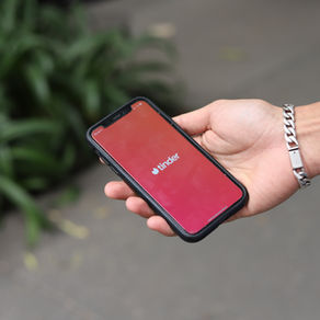 Hand hält Smartphone mit rotem Tinder-Startbildschirm; silbernes Armband, unscharfer grüner Hintergrund im Freien.