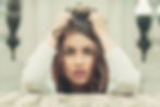 Woman with long hair grips head, looking intensely at the camera. Background is blurred, with stone and greenery. Mood is contemplative.