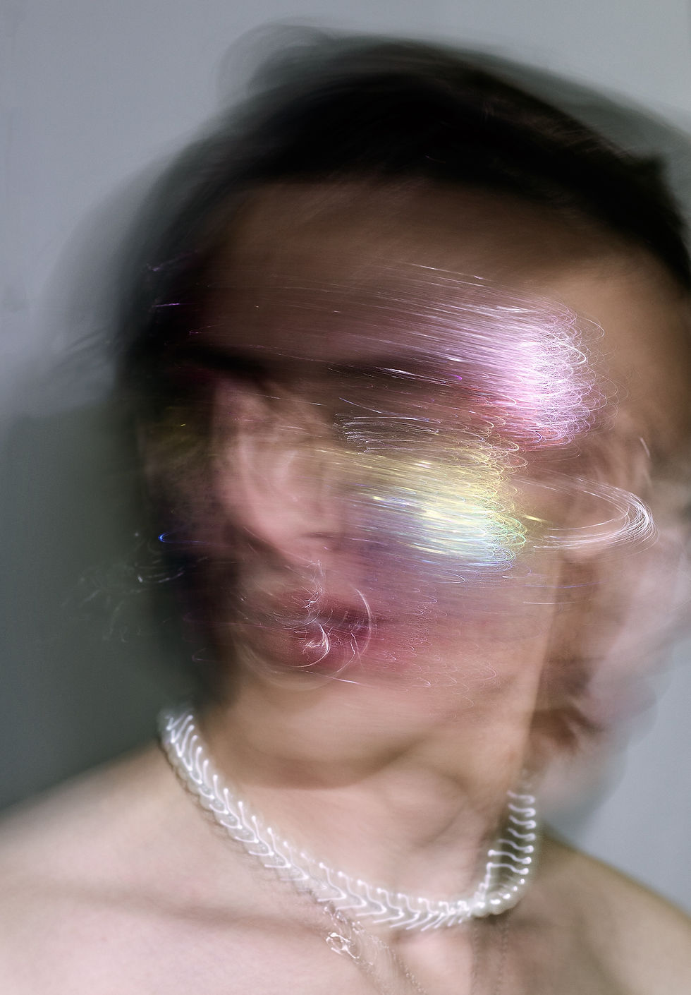 Blurry close-up of a person with swirling colorful light effects across their face. They wear a silver chain necklace against a plain background.
