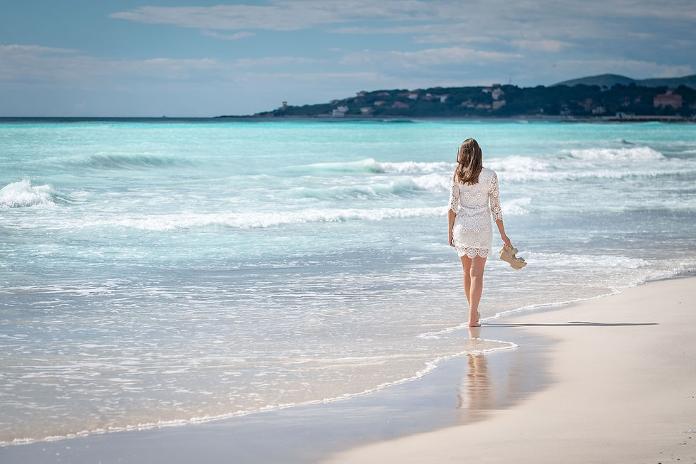 Femme en robe blanche marchant pieds nus sur une plage, tenant des sandales, face à la mer turquoise sous ciel bleu.