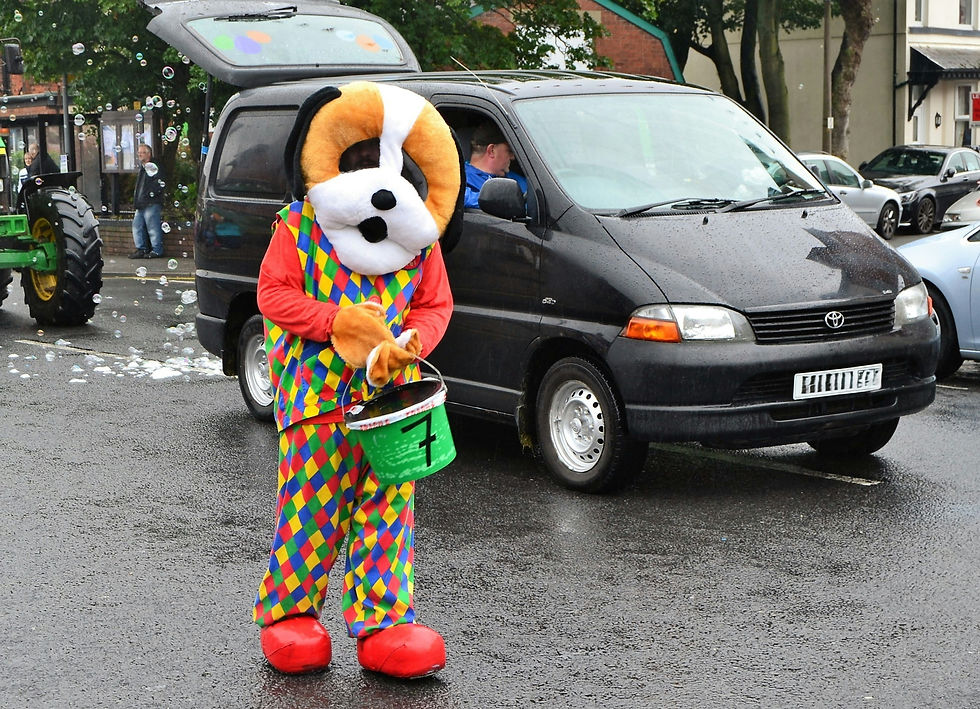 Person in a colourful dog mascot costume holds a green bucket on a rainy street beside a black van, with bubbles floating around.