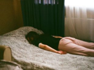 Person lying face down on a bed with a patterned cover, wearing pink pants and a black shirt. Room has dark curtains.
