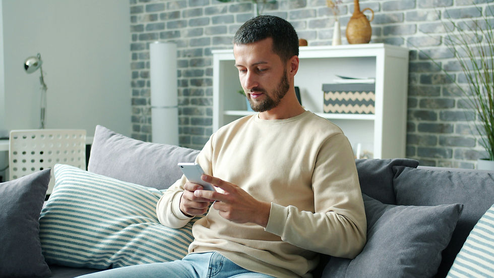 Homem sentado no sofá usa smartphone. Ambiente moderno com parede de tijolos, almofadas listradas em azul e prateleira com objetos decorativos.