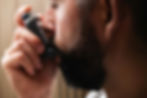 Man using a gray inhaler, close-up of hand and face. Neutral indoor setting with blurred brown background, focused on respiratory action.