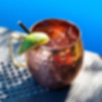 Copper mug with iced drink, lime wedge, and straw on a perforated table. Bright blue background, sunny, refreshing mood.