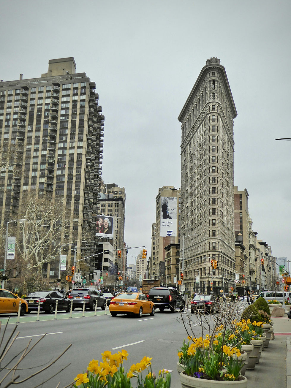 Overcast New York street with the Flatiron Building, traffic, and planters of yellow flowers lining the sidewalk.