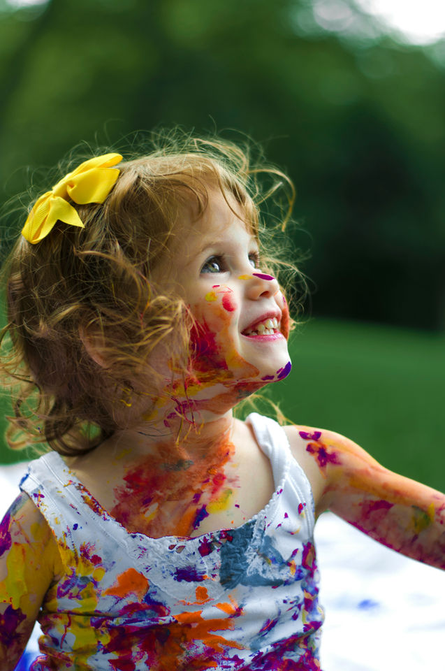 Smiling young girl covered in colorful paint, looking upwards in the yard.