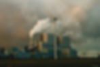 Factory with smoke stacks emitting steam; wind turbine in foreground. Cloudy sky; industrial, contrasting environment.