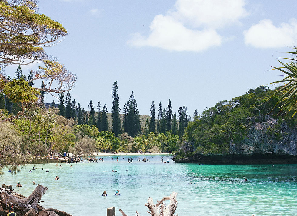 Swimmers in a turquoise tropical lagoon surrounded by lush trees, rocky cliffs, and a bright blue sky with clouds in New Caledonia