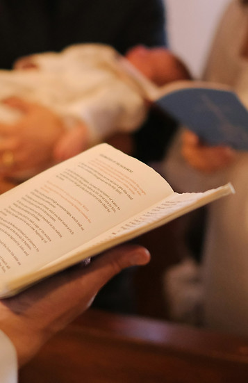 Bébé en robe de baptême blanche tenu par un parent tenant un livre religieux ouvert lors de la cérémonie de baptême.
