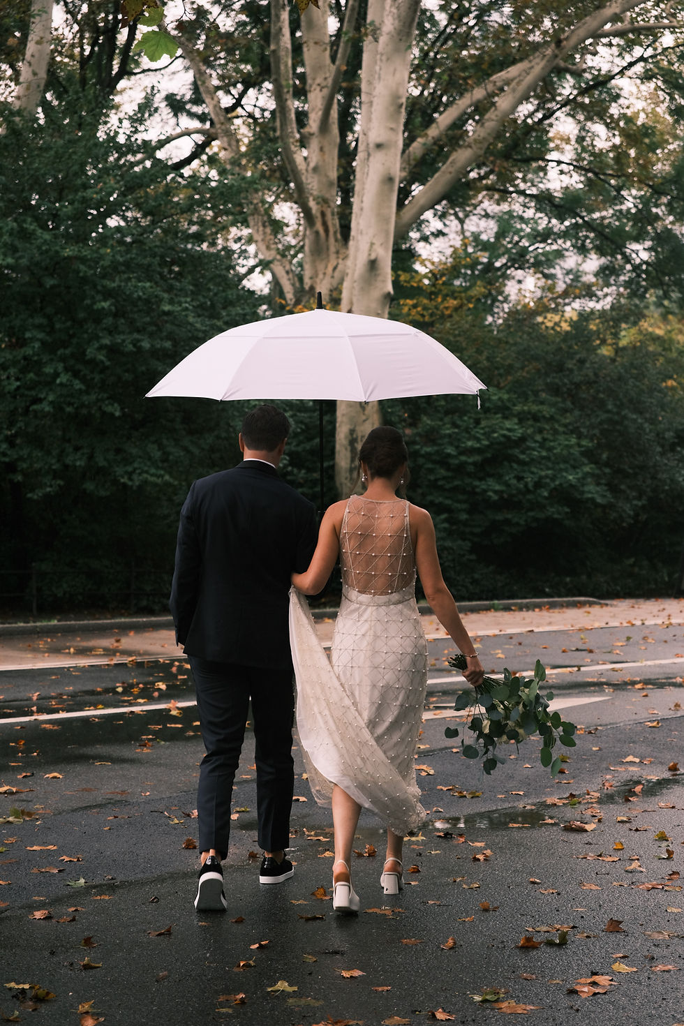 A newlywed couple strolls hand in hand under a white umbrella, enjoying a romantic walk on a rainy day surrounded by lush greenery.