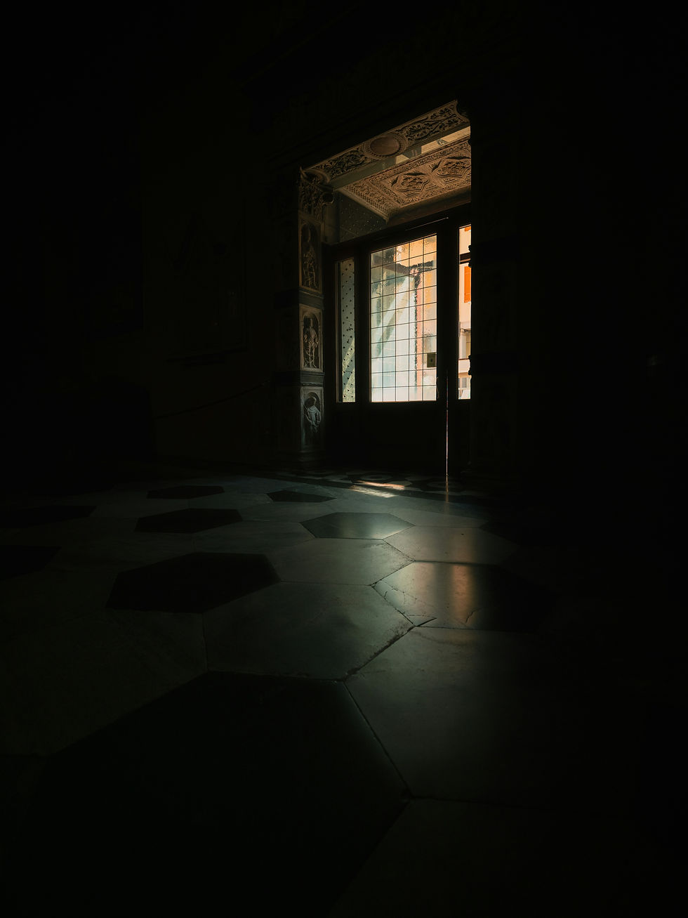Dim room with a partially open ornate door, letting in warm light onto hexagonal tiled floor. Dark, moody ambiance with decorative patterns.