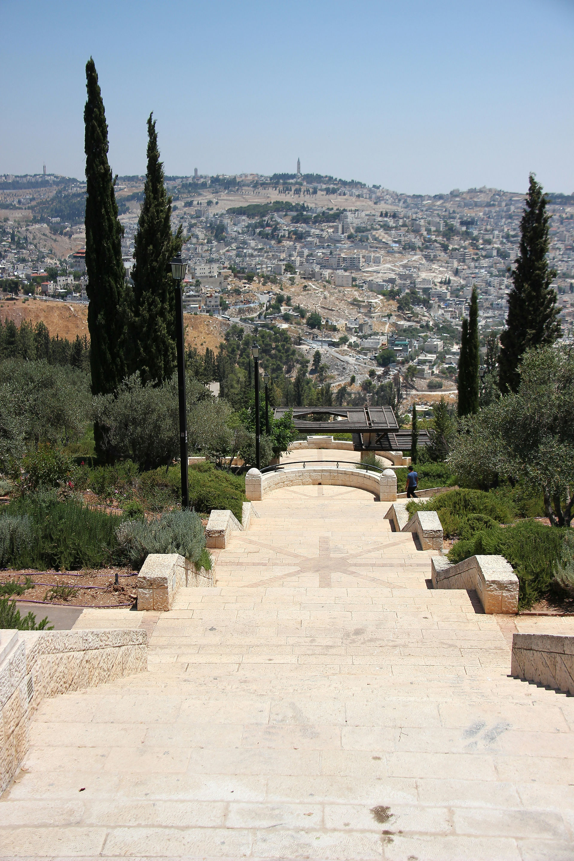Stone stairs leading down to the Kidron Valley from the Mount of Olives. A wide pedestrian path framed by cypress and olive trees with a view of the Silwan neighborhood.