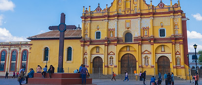 People walk and gather near a large stone cross in front of a vibrant yellow cathedral with ornate architectural details and arched doorways, set against a blue sky with scattered clouds.