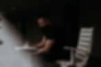 Man with a beard writing in a notebook, seated indoors on a white chair. Dark background with a soft light illuminating the scene.