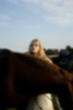 Blonde woman stands behind a brown horse in a grassy field under a pale blue sky, looking calm and thoughtful