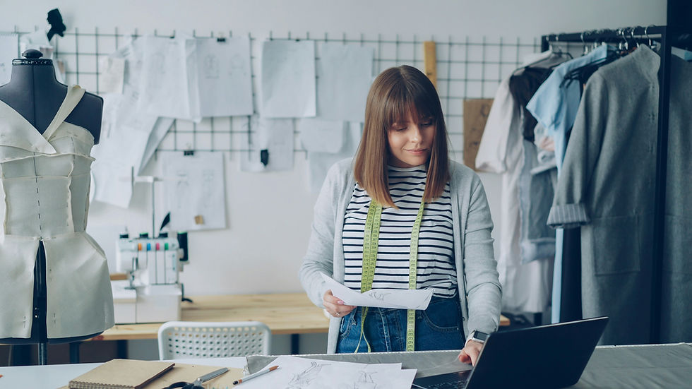 A woman in a striped shirt works at a desk with sewing patterns. Mannequin and sketches in background. Creative, focused atmosphere.