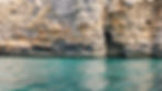 Layered tan cliff face above turquoise sea, with dark cave-like openings and calm rippling water below.