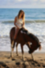 Young woman in white dress rides a brown horse on a sunny beach, waves behind her, calm seaside mood