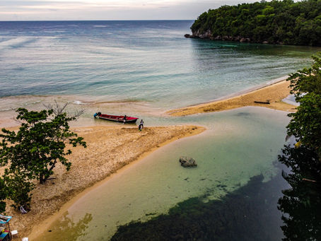 A red boat with two people rests on a tropical sandbar beside turquoise water, lush green cliffs, and calm sea.