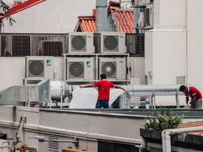 Two workers in red shirts install ducting on a rooftop beside Daikin AC units and vents, with white walls and orange-tiled roofs.
