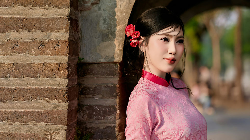 Mujer china con vestido rosa y flores rojas en el cabello, apoyada en un arco de ladrillo. Fondo natural desenfocado, expresión serena.