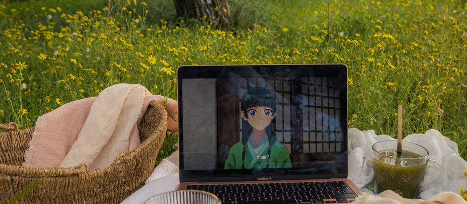 Laptop showing anime, surrounded by a picnic setup with croissants, flowers, and drinks on a white blanket under a tree in a field.