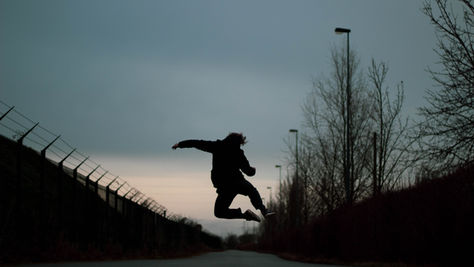 Silhouetted person mid-jump on an empty road at dusk, beside fence and bare trees under a gray sky