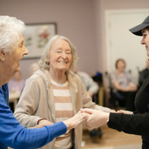 Elderly women joyfully hold hands with a caregiver in an assisted living facility, creating a warm, friendly atmosphere. The caregiver wears a cap with "SCCH."