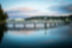 Tranquil harbor scene of Bainbridge Island with a wooden pier over calm water, boats docked, and a ferry in the distance. Soft pastel clouds in the sky.