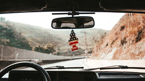 View from inside a car on a winding mountain road, with a US flag tree air freshener hanging below the rearview mirror.